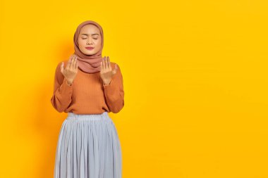 Kahverengi süveter ve tesettürlü güzel bir Asyalı kadın, elleri dua jesti içinde katlanmış, sarı arka planda izole edilmiş bir şey için yalvarıyor. İnsanlar İslam dini kavramları