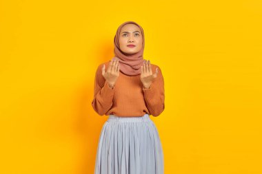 Kahverengi süveter ve tesettürlü güzel bir Asyalı kadın, elleri dua jesti içinde katlanmış, sarı arka planda izole edilmiş bir şey için yalvarıyor. İnsanlar İslam dini kavramları