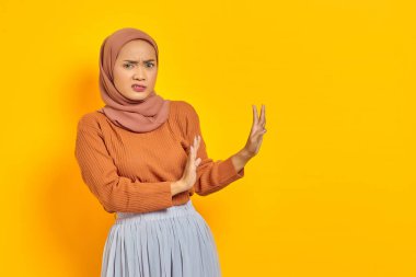 Kahverengi kazaklı ve tesettürlü güzel Asyalı kadın dur hareketi yapıyor, sarı arka planda izole edilmiş reddedilmeyi gösteriyor. İnsanlar dini yaşam tarzı konsepti