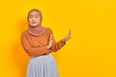 Kahverengi kazaklı ve tesettürlü güzel Asyalı kadın dur hareketi yapıyor, sarı arka planda izole edilmiş reddedilmeyi gösteriyor. İnsanlar dini yaşam tarzı konsepti