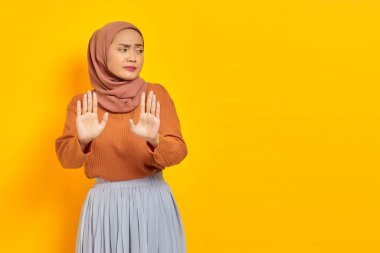 Kahverengi kazaklı ve tesettürlü güzel Asyalı kadın dur hareketi yapıyor, sarı arka planda izole edilmiş reddedilmeyi gösteriyor. İnsanlar dini yaşam tarzı konsepti