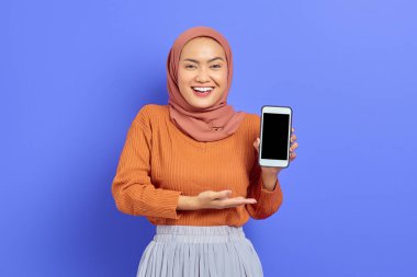 Kahverengi kazak ve tesettürlü gülümseyen Asyalı güzel bir kadın mor arka planda izole edilmiş boş ekran cep telefonu gösteriyor. İnsanlar İslam dini kavramları