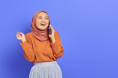 Kahverengi süveter ve tesettürlü neşeli Asyalı kadın mor arka plandan izole edilmiş bir tarafa bakarken cep telefonuyla konuşuyor. İnsanlar İslam dini kavramları