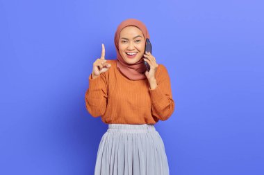 Kahverengi kazaklı, tesettürlü ve tesettürlü güzel bir Asyalı kadının portresi. Elinde cep telefonu olan ve mor arka planda izole edilmiş bir parmak gösteren. İnsanlar İslam dini kavramları