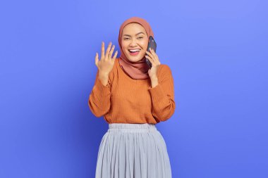 Kahverengi süveter ve tesettürlü güzel bir Asyalı kadın cep telefonuyla konuşurken mor arka planda izole edilmiş avuç içlerini gösteriyor. İnsanlar İslam dini kavramları