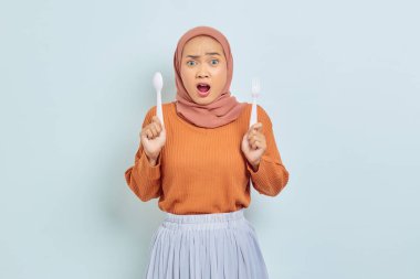 Kahverengi kazak ve tesettürlü güzel Asyalı kadın beyaz arka planda yalnız kahvaltı etmeden önce çatal ve kaşık tutuyor.