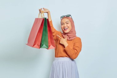 Kahverengi kazaklı ve gözlüklü neşeli Asyalı Müslüman kadın beyaz arka planda parmak izole edilmiş bir alışveriş torbasını işaret ediyor. Dünya Tüketici Hakları Günü Kavramı