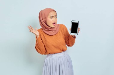 Kahverengi kazak ve tesettürlü güzel bir Asyalı kadın, beyaz arka planda ağzı açık, boş bir cep telefonu ekranı gösteriyor. Müslüman yaşam tarzı kavramı