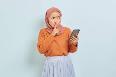 Kahverengi kazaklı ve tesettürlü güzel bir Asyalı kadın akıllı telefon tutuyor, parmakları dudaklarını kapatıyor, beyaz arka planda izole edilmiş sessiz bir jest yapıyor. Müslüman yaşam tarzı kavramı