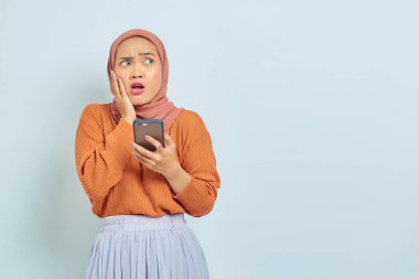 Kahverengi kazaklı, cep telefonu kullanan ve beyaz arka planda izole edilmiş ağzı açık bir Asyalı genç Müslüman kadın. Müslüman yaşam tarzı kavramı
