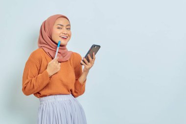 Kahverengi kazaklı tesettürlü gülümseyen Asyalı Müslüman kadının portresi ve beyaz arka planda izole edilmiş cep telefonu kullanımı. Müslüman yaşam tarzı kavramı