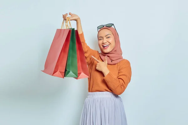 身穿棕色毛衣和眼镜的快乐美丽的亚洲穆斯林妇女指着购物袋 手指与白色背景隔离 世界消费者权利日概念 — 图库照片
