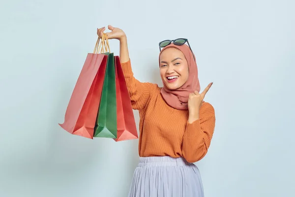 穿着棕色毛衣 戴着眼镜 手里拿着购物袋的快乐美丽的亚裔穆斯林女人 同时用手指在白色背景上复制空间 世界消费者权利日概念 — 图库照片