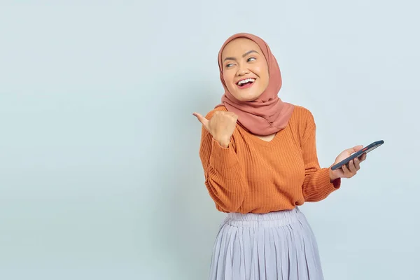 Веселая Красивая Азиатская Мусульманка Коричневом Свитере Хиджабе Держащая Смартфон Указывая — стоковое фото