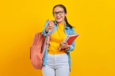 Sırt çantalı, sırt çantalı, sarı arka planda kamerayı işaret eden heyecanlı Asyalı kadın öğrenci portresi. Lise üniversite konsepti eğitimi