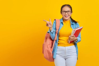 Sırt çantalı ve sırt çantalı gülümseyen Asyalı genç üniversite öğrencisi portresi sarı arka planda izole edilmiş kopya alanını işaret ediyor.
