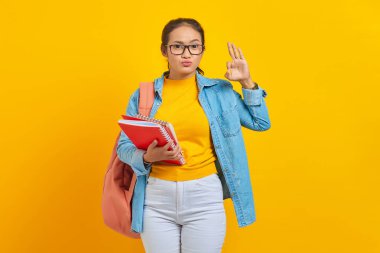 Sırt çantalı, kot elbiseli, elinde not defteri olan ve sarı arka planda izole edilmiş iyi bir hareket gösteren komik genç bayan öğrenci. Lise üniversite konsepti eğitimi
