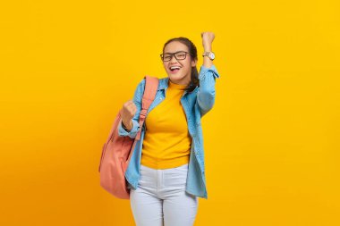 Sırt çantalı neşeli Asyalı öğrenci sarı arka planda izole edilmiş evet diyerek birinci sınıf jest yapıyor. Lise üniversite konsepti eğitimi