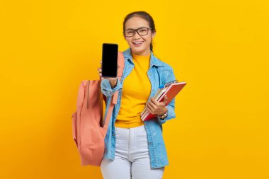 Sırt çantası taşıyan ve sarı arka planda izole edilmiş boş ekran cep telefonunu gösteren kot pantolon giymiş gülümseyen Asyalı genç kadın portresi.