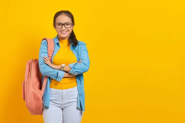 Smiling young Asian student in denim outfit with backpack crossed chest and looking confident isolated on yellow background. Education in high school university college concept