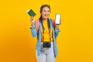 Pasaport kartı gösteren neşeli Asyalı genç bir gezginin portresi ve sarı arka planda izole edilmiş boş ekran cep telefonu. Hafta sonları seyahat eden yolcular. Uçak yolculuğu kavramı