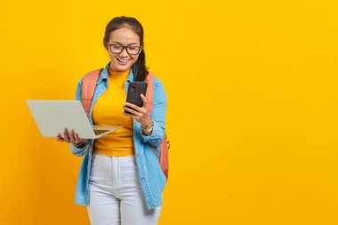 Sırt çantalı, laptoplu ve sarı arka planda izole edilmiş akıllı telefondan haber mesajı okuyan neşeli Asyalı kız öğrenci. Üniversite konsepti eğitimi