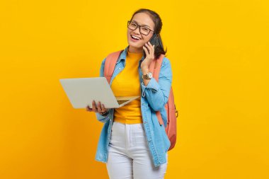 Sırt çantalı, cep telefonuyla konuşan ve sarı arka planda dizüstü bilgisayar tutan neşeli Asyalı genç kız öğrenci portresi. Üniversite konsepti eğitimi