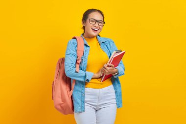 Sırt çantasında kitap taşıyan ve sarı arka planda izole edilmiş kameraya bakan sıradan giyinmiş neşeli Asyalı genç kadın portresi.