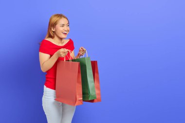 Kırmızı elbiseli neşeli, güzel Asyalı kadın alışveriş çantası açıyor ve mor arka planda soyutlanmış bir kenara bakıyor.