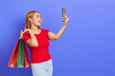 Kırmızı elbiseli neşeli güzel Asyalı kadın alışveriş torbaları tutuyor ve mor arka planda izole edilmiş cep telefonundan selfie çekiyor.