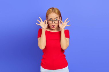 Şaşırmış Asyalı bir kadının portresi. Elinde gözlük, ağzı açık ve önüne bakıyor. Mor arka planda soyutlanmış.