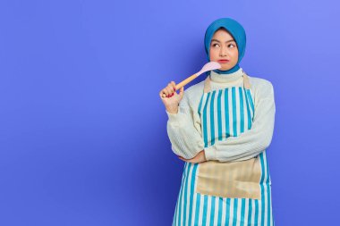 Beyaz önlük ve tesettüre bürünmüş güzel Asyalı kadın elinde kaşık, mor arka planda izole edilmiş ev işlerini yapıyor. Oda servisi kavramı
