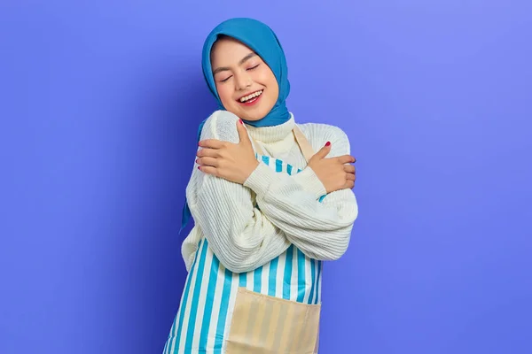 穿着头巾和条纹围裙的年轻的亚洲穆斯林妇女的画像 拥抱自己 觉得冷 被紫色背景隔离 人们家庭妇女穆斯林生活方式的概念 — 图库照片