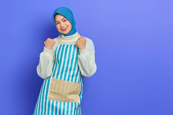 身穿头巾和围裙的快乐的年轻的亚洲穆斯林妇女 庆祝成功 并得到了与紫色背景隔离的好消息 人们家庭妇女穆斯林生活方式的概念 — 图库照片