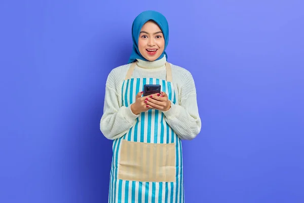 紫色の背景に隔離されたカメラを見ながら 携帯電話を使ってヒジャーブとエプロンを身に着けている20代の陽気なアジア系ムスリム女性の肖像画 主婦ムスリムライフスタイルの概念 — ストック写真