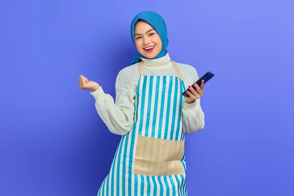 대젊은 아시아 히잡을 앞치마를 핸드폰을 보라색 배경에서 승자의 몸짓을 흥분된 — 스톡 사진