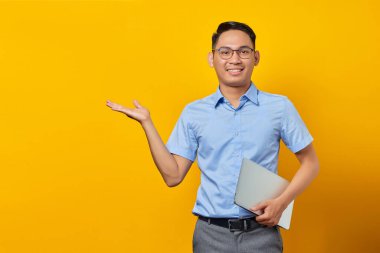 Gözlüklü gülümseyen Asyalı adamın portresi dizüstü bilgisayarlı ve sarı arka planda avuç içi izole edilmiş fotokopi alanını gösteriyor. İş adamı ve girişimci konsepti