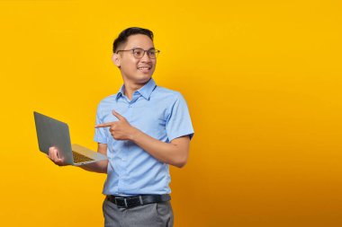 Gözlüklü yakışıklı Asyalı adamın portresi dizüstü bilgisayarı işaret ediyor, sarı arka planda internetten izole edilmiş bir şey gösteriyor. İş adamı ve girişimci konsepti