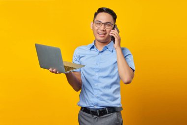 Gözlüklü neşeli Asyalı genç adam cep telefonuyla konuşuyor ve sarı arka planda dizüstü bilgisayar kullanıyor. İş adamı ve girişimci konsepti