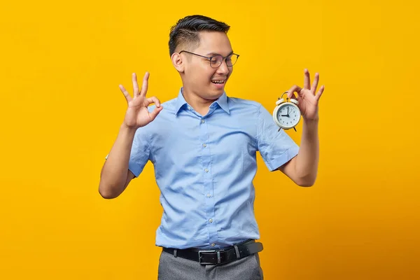 一副笑容可亲 英俊的亚洲年轻人的画像 戴着一副眼镜 显示出闹钟的样子 用黄色背景隔离的手指 表现出一副不错的姿势 时间管理生活方式概念 — 图库照片