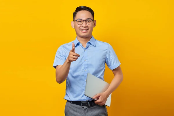 身穿眼镜 手持笔记本电脑 用手指指向被黄色背景隔离的相机的英俊的亚洲男子的画像 商人和企业家概念 — 图库照片