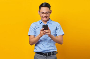 Gözlüklü gülümseyen Asyalı genç adam sarı arka planda izole edilmiş cep telefonundan gelen mesajlara cevap veriyor. İş adamı ve girişimci konsepti