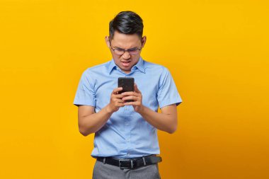 Gözlüklü Asyalı üzgün genç adam sarı arka planda izole edilmiş cep telefonundan gelen mesajlara cevap veriyor. İş adamı ve girişimci konsepti