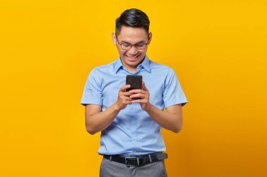 Gözlüklü, gülen Asyalı genç adam sarı arka planda izole edilmiş cep telefonundan gelen mesajlara cevap veriyor. İş adamı ve girişimci konsepti