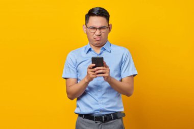 Gözlüklü dalgın Asyalı genç adam sarı arka planda izole edilmiş cep telefonundan gelen mesajlara cevap veriyor. İş adamı ve girişimci konsepti