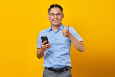 Klasik mavi gömlekli, gülümseyen genç, yakışıklı iş adamı cep telefonu kullanıyor ve sarı arka planda izole edilmiş baş parmak işareti yapıyor. Kariyer zenginliği iş konseptini elde etmek