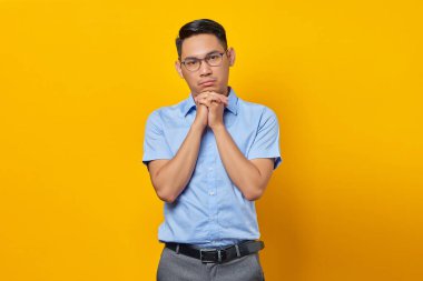 Gözlüklü düşünceli genç Asyalı adamın portresi sarı arka planda izole edilmiş bir çenenin altında el ele tutuyor. İş adamı ve girişimci konsepti