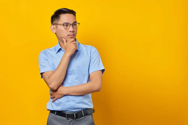 戴眼镜沉思的年轻亚洲男子的画像 双手插在下巴下 黄色背景隔离 商人和企业家概念 — 图库照片