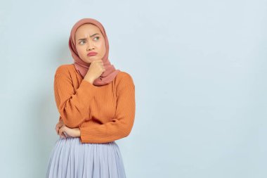 Kahverengi kazaklı, Asyalı genç bir Müslüman kadının portresi. Soruyu ciddi ciddi düşünüyor ve beyaz arka planda izole edilmiş fotokopi alanına bakıyor. Müslüman yaşam tarzı kavramı