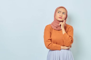 Kahverengi kazaklı, Asyalı genç bir Müslüman kadının portresi. Soruyu ciddi ciddi düşünüyor ve beyaz arka planda izole edilmiş fotokopi alanına bakıyor. Müslüman yaşam tarzı kavramı